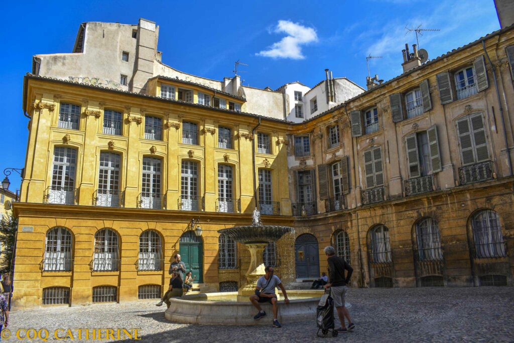 La Place d’Albertas d’Aix en Provence avec des façades ocres et une fontaine au centres avec des personnes autour