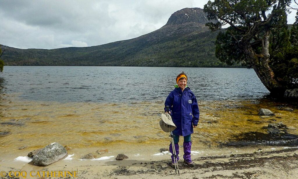Kat devant un lac avec une veste de pluie, un chapeau et des guêtres