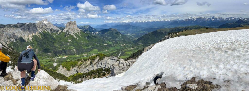 Panorama sur les montagnes des Ecrins, le Dévoluy et l’Obiou depuis le sommet de la Tête Chevalière dans le Vercors, avec un plateau herbeux, un grand névé et les falaises calcaires